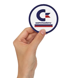 Può includere: Un cerotto rotondo bianco con un bordo blu e un logo "C" rosso e blu. Il testo "commodore" è scritto in blu sotto il logo.