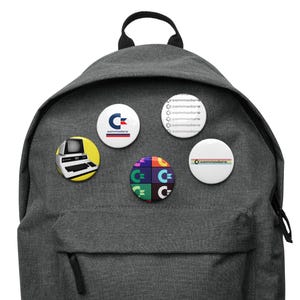 May include: A gray backpack with five different Commodore-themed buttons. The buttons feature a retro computer, a logo with the word "Commodore" and a rainbow stripe.