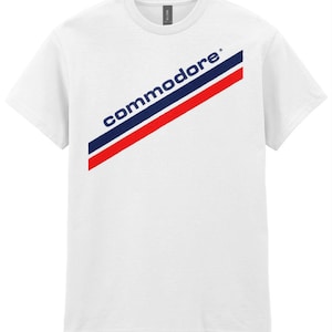 Könnte beinhalten: Weißes T-Shirt mit einem diagonalen rot-weiß-blauen Streifen und dem Wort "commodore" in blauen Buchstaben.