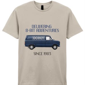 Könnte beinhalten: Beiges T-Shirt mit einer dunkelblauen Van-Grafik. Der Van hat das Wort "ocean" auf der Seite. Der Text "DELIVERING 8-BIT ADVENTURES" und "SINCE 1983" befinden sich über und unter der Van-Illustration.