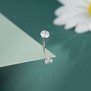 Può includere: Un piercing per l'ombelico curvo con una barra in metallo color argento. Ogni estremità dell'anello presenta una gemma trasparente, rotonda e scintillante. Il gioiello è posto su uno sfondo verde acqua e bianco.