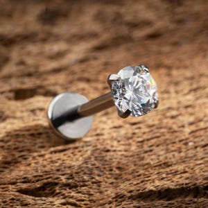 May include: A silver-toned labret stud body jewelry piece featuring a round, clear, faceted gemstone held in a four-prong setting. The post is straight and connects to a flat, circular disc base, designed for various piercings.
