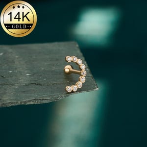 May include: A gold-colored, curved earring with a series of small, clear gemstones. The earring is set against a dark, textured surface, with a gold-colored emblem in the upper left corner that reads "14K GOLD".