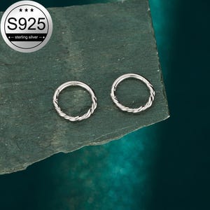 May include: Two silver-colored hoop earrings with a twisted rope design. The earrings are displayed on a dark green, textured surface. The image also includes a circular logo that reads "S925 - sterling silver".