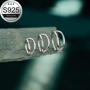 Könnte beinhalten: Drei silberne Creolen in verschiedenen Größen, jede mit einem Doppelring-Design. Ein kreisförmiges Emblem in der oberen linken Ecke zeigt den Text S925 und sterling silver, was die Materialzusammensetzung der Schmuckstücke angibt.