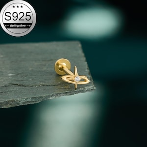 Peut inclure: Boucle d'oreille dorée avec un motif en forme d'éclat d'étoile et une petite pierre précieuse transparente au centre. La boucle d'oreille est en argent sterling S925. La boucle d'oreille est sur une surface gris foncé.