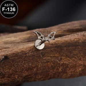 May include: A silver-toned titanium body jewelry stud featuring a detailed bat-shaped top with spread wings. The piece includes a flat circular back disc and is labeled with the text ASTM F-136 TITANIUM, indicating the material grade used.