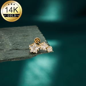 Puede incluir: Un pendiente dorado con tres gemas claras en forma de estrella. El pendiente está engarzado en un poste y cierre dorado. La imagen también incluye un emblema dorado que dice "14K GOLD".