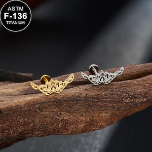 Op de afbeelding: Twee ASTM F-136 titanium lichaamsieraden met een lotusbloemontwerp. Het ene sieraad is goudkleurig en het andere is zilverkleurig. Elke stud heeft een platte achterkant en een decoratieve voorkant in de vorm van een open, symmetrische lotusbloem.