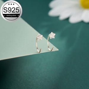 May include: A pair of silver hoop earrings, each with a flower-shaped top. The earrings are made of S925 sterling silver. They are displayed on a light green surface against a dark green background. The earrings are a delicate jewellery item.
