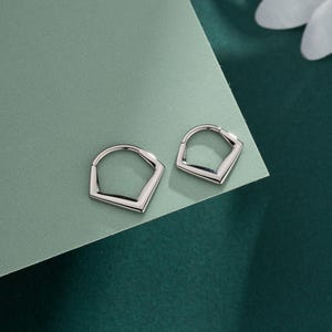 Könnte beinhalten: Ein Paar silberfarbene Creolen. Jeder Ohrring hat eine geometrische, pentagonartige Form mit gewölbter Oberseite und geradem Boden. Die Ohrringe werden auf einer grünen Oberfläche präsentiert.
