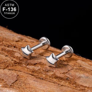 May include: Two silver-colored star-shaped flat back labret studs made of ASTM F-136 titanium. Each piece features a polished star top attached to a straight post with a circular flat base, designed for body piercings and jewelry applications.
