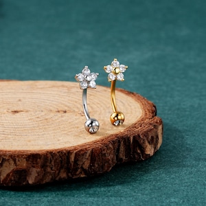Puede incluir: Dos piezas de joyería corporal de barra curva, una en plata y otra en oro, con un diseño de flor engastada con piedras de circonita cúbica transparentes.