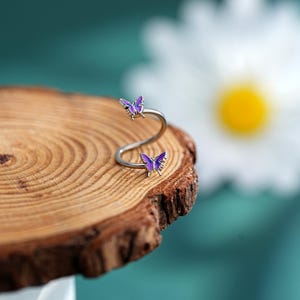 May include: A silver-toned ring with a swirling design, featuring two purple butterfly accents. The ring is set against a wooden surface with a blurred background, showcasing a daisy.
