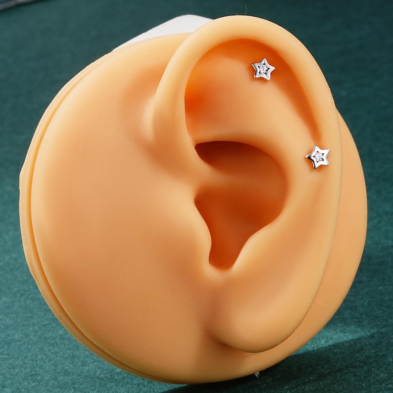 May include: Two small silver star-shaped earrings with clear cubic zirconia stones set in the center of each star. The earrings are shown on a model ear.