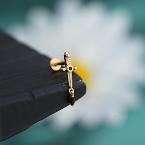 Puede incluir: Pendiente dorado en forma de espada con una gema negra en el centro y una gema negra colgante en forma de lágrima. El pendiente está colocado sobre una superficie oscura con un fondo blanco y verde borroso.