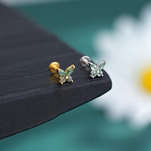 May include: Two butterfly stud earrings. One has a gold post with a butterfly design featuring green and clear stones. The other has a silver post with a similar butterfly design. The earrings are on a black surface.