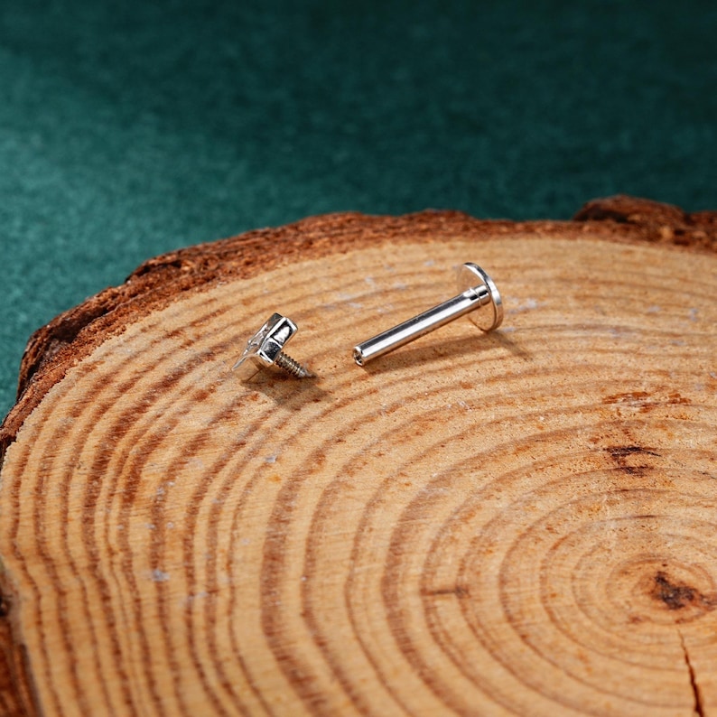 May include: A silver body jewelry piercing with a flat back and a screw-on top. The jewelry is on a wooden surface.