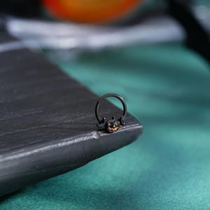 May include: Black nose ring featuring a small, orange and black jack-o'-lantern charm. The ring is a circular design with a curved bar and a pumpkin pendant. The jewelry is likely for Halloween or fall-themed outfits.