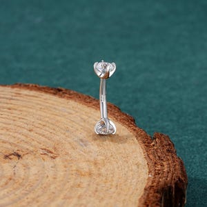 May include: A silver body piercing with two clear crystal stones. The piercing is curved and designed for a belly button.