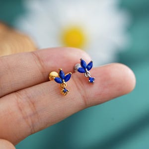 Op de afbeelding: Een paar gouden en zilveren oorstekers met een bloemmotief. Elke oorbel heeft een cluster van blauwe marquise-geslepen edelstenen en een kleine vierkante edelsteen. De oorbellen worden op een lichtgekleurde achtergrond getoond.