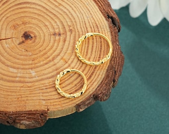 May include: Two gold-colored hoop earrings with a braided design. The earrings are displayed on a wooden surface with a natural wood grain pattern. The background is a dark teal color, with a white flower in the upper right corner.