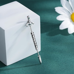 May include: A silver-colored sword-shaped earring with a detailed handle and blade design. The earring is displayed on a white block against a green background, with a daisy in the background.