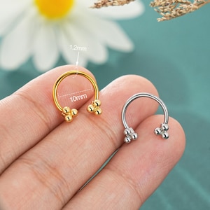 May include: A gold-colored septum ring with a 10mm diameter and 1.2mm thickness. The ring features three small, round decorative elements on each end. A silver-colored septum ring is also visible.