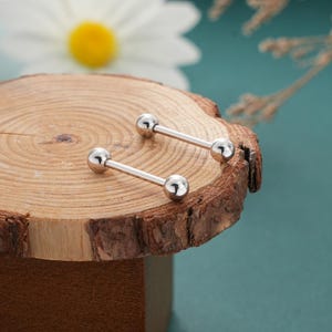 May include: Two silver barbell earrings with a ball on each end, displayed on a wooden surface. The earrings are shiny and reflect light. The background includes a daisy and a teal surface.