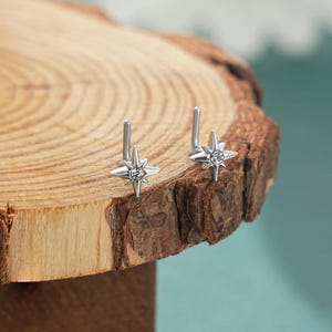 Puede incluir: Dos piercings de nariz plateados, cada uno con un diseño de estrella con una gema clara central. Los piercings se muestran sobre una superficie de madera rústica, con un fondo azul suave.