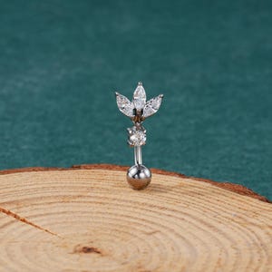 Puede incluir: Un piercing de ombligo plateado con un diseño de flor de cristal transparente.