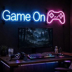 Op de afbeelding: Een neonbord aan de muur met de tekst Game On in blauw en een roze icoon van een gamecontroller. Het bord hangt boven een computermonitor, een mechanisch toetsenbord met RGB-verlichting, een computermuis en een headset op een houten bureau.