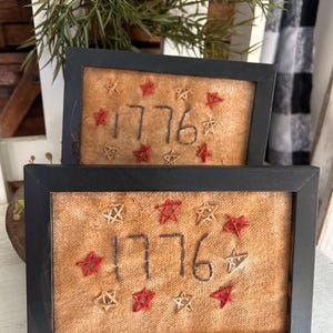 May include: Two framed pieces of art featuring the number "1776" stitched in dark thread, surrounded by red and tan star embroidery. The frames are black and rectangular, with a rustic, vintage aesthetic.