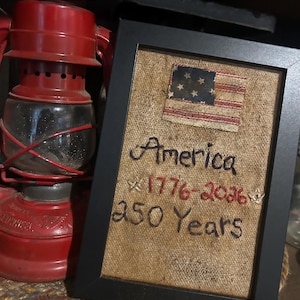 May include: A framed burlap artwork with a small American flag patch. The text reads "America 1776-2026 250 Years" in black and red thread. A red lantern is visible in the background, suggesting a rustic or patriotic theme.