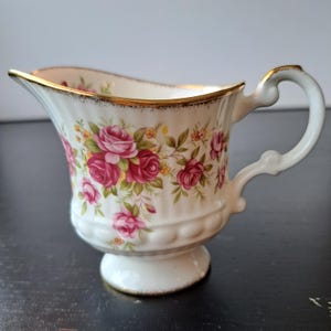 May include: A white porcelain creamer featuring a fluted body, a pedestal base, and a curved handle. The exterior is decorated with a pattern of pink roses, green leaves, and small yellow flowers, finished with a gold trim along the rim and base.