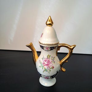 Op de afbeelding: Een porseleinen theepot met een crèmekleurig lichaam en een bloemmotief van roze rozen en groene bladeren. De theepot heeft een goudkleurig handvat, tuit en dekselknop, geaccentueerd door donkerblauwe decoratieve banden rond de hals en de voet.