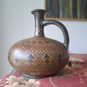 Può includere: Una brocca in ceramica marrone con corpo arrotondato e manico. La brocca presenta intricati motivi geometrici e di uccelli in nero e rosso. La brocca ha collo e manico marrone scuro e un piccolo tappo di legno.