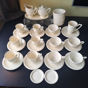 Könnte beinhalten: Ein cremefarbenes Teeservice mit mehreren Tassen, Untertassen, Schalen, einem Sahnekännchen und einer Zuckerdose. Das Set ist auf einer dunklen Oberfläche angeordnet. Die Tassen und Untertassen haben ein schlichtes, elegantes Design.