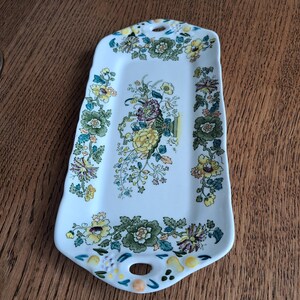 Op de afbeelding: Een rechthoekig wit keramieken serveerblad met geschulpte randen en twee geïntegreerde handgrepen. Het oppervlak is voorzien van een centraal bloemstuk in geel, groen en paars, omringd door een doorlopende rand met botanische patronen.