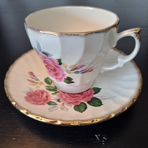 Könnte beinhalten: Ein weißes Teetassen- und Untertassenset aus Porzellan mit einem zarten rosa Rosenmuster, grünen Blättern und Knospen. Der Rand der Tasse und der Untertasse ist mit einem goldenen Metallrand verziert, und die Tasse weist ein geriffeltes, gewelltes Design auf.