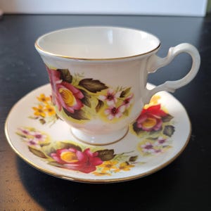 May include: A white porcelain teacup and saucer set featuring a floral pattern of red roses, yellow wildflowers, and small pink blossoms. The rim of both the cup and the saucer is accented with a thin gold trim, and the cup has a curved handle.
