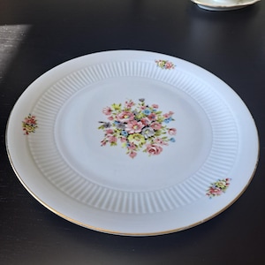 Op de afbeelding: Een wit porseleinen bord met een centraal bloemenboeket van roze, gele en blauwe bloemen. De rand heeft een geribbelde textuur en is versierd met drie kleinere bloemmotieven. De rand is afgewerkt met een dunne gouden bies.