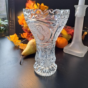 Op de afbeelding: Een heldere, sierlijke glazen vaas met een geschulpte rand en een gedetailleerd paisley-ontwerp. De vaas heeft een uitlopende vorm en staat op een decoratieve voet. De achtergrond bevat herfstbladeren en een matglazen kandelaar.