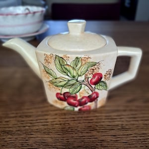 Könnte beinhalten: Eine cremefarbene Keramik-Teekanne mit einem handgemalten Motiv aus roten Kirschen an einem Zweig mit grünen Blättern. Die Teekanne hat einen quadratischen Körper, einen passenden Deckel mit Knauf, einen Ausguss und einen stabilen Henkel.