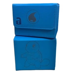 May include: Two-piece blue card storage box. The top box has a flap closure with a grey logo and a keyhole design. The bottom box features a Squirtle character design. Both boxes are made of a textured material.