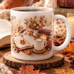 May include: A white ceramic mug featuring a printed scene of an open book, a small cup of coffee, a stack of books, a lit candle, and a vase of autumn flowers, all surrounded by scattered maple leaves and a cozy knitted texture.