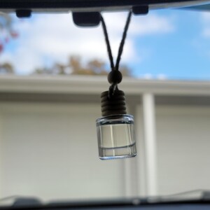May include: A clear, rectangular car air freshener bottle with a dark cap and a black string for hanging. The bottle is filled with a clear liquid and hangs from a car's rearview mirror, with a blurred background of a blue sky and a house.