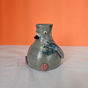 May include: A ceramic pigeon-shaped vase. The vase is grey with blue wing details and pink feet. It has a black beak, white face, and a single eye. The background is a solid orange.