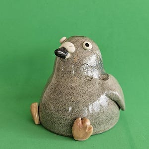 May include: A ceramic bird ornament with a speckled grey body, black beak, and small, round eyes. The bird has small, tan feet and is set against a solid green background. The ornament has a glossy finish.