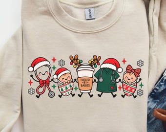 May include: A beige crewneck sweatshirt featuring a graphic of a stethoscope, a swaddled baby, a coffee cup with reindeer antlers, a scrub top, and another swaddled baby, all wearing Christmas accessories. The coffee cup has the text FUELED BY BABIES.